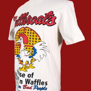May include: White t-shirt featuring a cartoon rooster with a red comb and yellow feet, standing in front of a waffle. The shirt has the text "Fullthroats" in red and the words "House of Chicken and Waffles for Bad People".