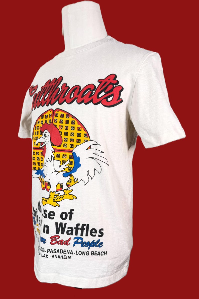May include: White t-shirt featuring a cartoon rooster with a red comb and yellow feet, standing in front of a waffle. The shirt has the text "Fullthroats" in red and the words "House of Chicken and Waffles for Bad People".