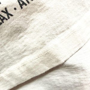 May include: Close-up of a light gray t-shirt with black text that reads "LAX ANAH". The fabric has a textured appearance with visible stitching along the seams. The shirt appears to be made of a soft, breathable material.