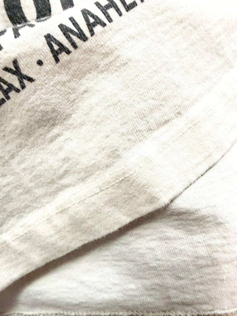 May include: Close-up of a light gray t-shirt with black text that reads "LAX ANAH". The fabric has a textured appearance with visible stitching along the seams. The shirt appears to be made of a soft, breathable material.