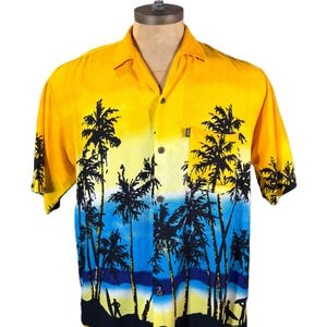 May include: Yellow, blue, and black button-down shirt with a tropical beach scene. The shirt features a gradient design, transitioning from yellow to blue, with black palm trees and silhouettes of surfers. It has a pocket on the chest.
