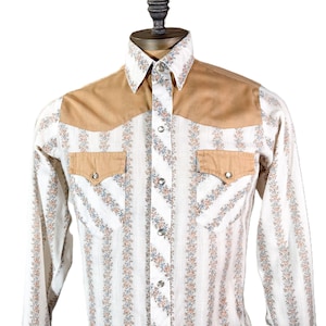 May include: A vintage Western-style shirt with a white background and vertical floral stripes in blue, orange, and green. The shirt features tan suede-like shoulder and pocket accents, snap closures, and a pointed collar. The long-sleeved shirt is displayed on a mannequin.