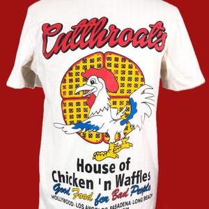 May include: White t-shirt with a graphic of a rooster and the words "Cutthroats" in red script. Below the rooster is the text "House of Chicken 'n Waffles" and the phrase "Good Food for Bad People."