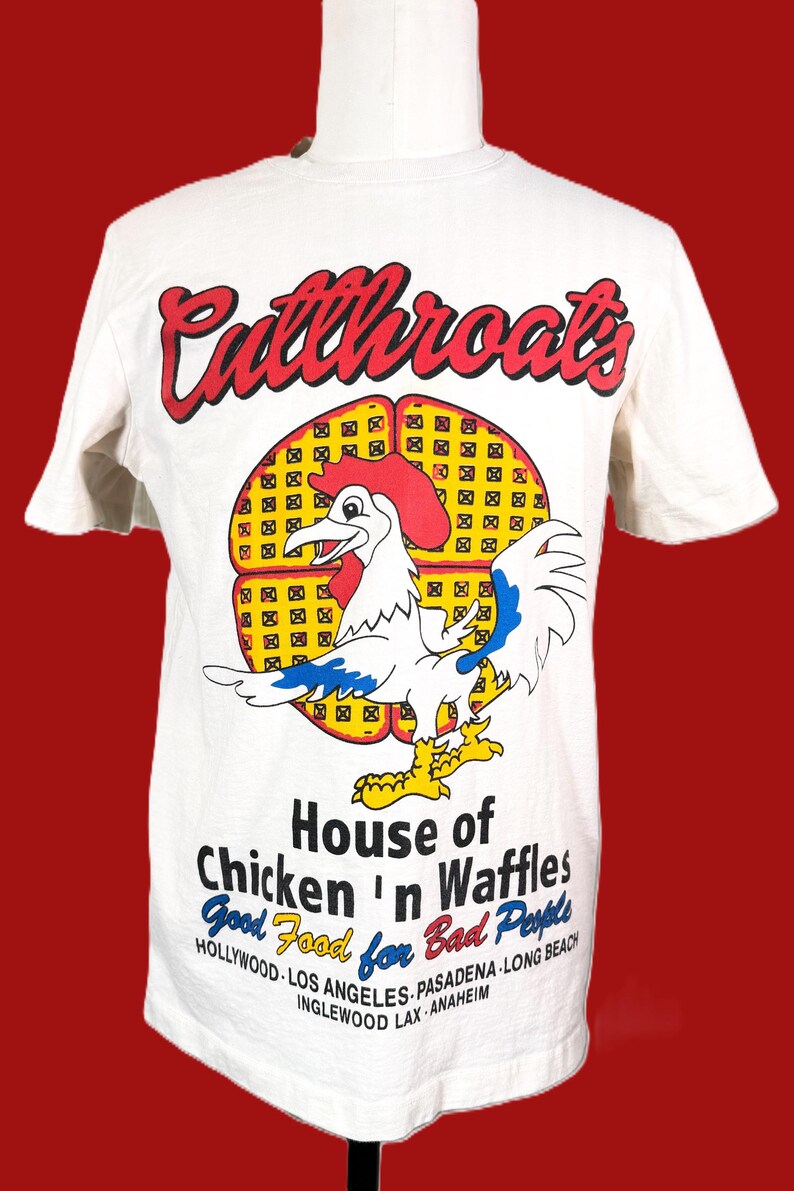 May include: White t-shirt with a graphic of a rooster and the words "Cutthroats" in red script. Below the rooster is the text "House of Chicken 'n Waffles" and the phrase "Good Food for Bad People."