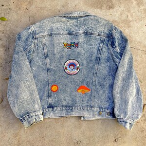 May include: Acid wash denim jacket with colorful embroidered patches. The patches include a dancing bear design, a circular patch with a woman's face, a smiling sun, and a mushroom. The jacket has a button-down front and two chest pockets.