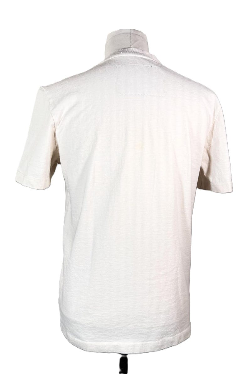 May include: A cream-colored short-sleeved t-shirt displayed on a mannequin. The shirt has a crew neck and a slightly textured appearance. The back of the shirt is visible, showing the simple design.