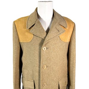 May include: A tan tweed jacket with tan suede shoulder patches. The jacket has a notched collar, three buttons, and two front pockets. The jacket is displayed on a white mannequin.