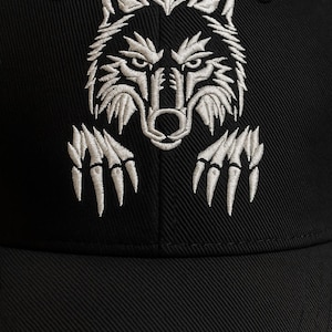 Puede incluir: Gorra de béisbol negra con un diseño de lobo bordado en blanco. La cabeza y las garras del lobo se muestran de forma prominente, creando un contraste llamativo con la tela oscura. La gorra es de estilo clásico, adecuada para uso casual.