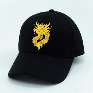 May include: Black baseball cap featuring a gold embroidered dragon design. The dragon has red eyes and intricate detailing. The cap has a curved brim and a button on top, suitable for casual wear.