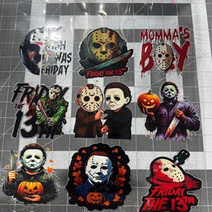 May include: A collection of nine horror-themed stickers featuring Jason Voorhees and Michael Myers. Designs include hockey masks, pumpkins, and text such as "Momma's Boy" and "Friday the 13th." The stickers are arranged on a cutting mat with inch markings.