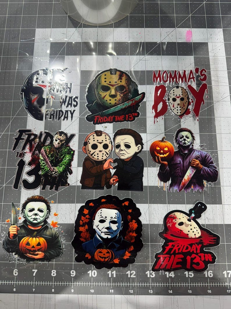May include: A collection of nine horror-themed stickers featuring Jason Voorhees and Michael Myers. Designs include hockey masks, pumpkins, and text such as "Momma's Boy" and "Friday the 13th." The stickers are arranged on a cutting mat with inch markings.