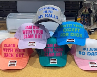 May include: A collection of six trucker hats in various colors including neon orange, lavender, teal, blue, and pink. Each hat features a foam front panel with bold text printed in contrasting colors, including phrases like SCISSORING HURT MY BALLS and ALL SUGAR NO DADDY.