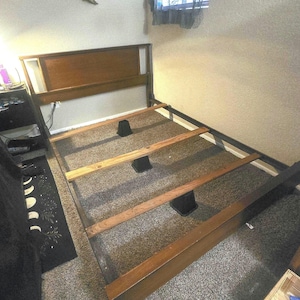 May include: A wooden bed frame featuring a rectangular headboard with a smooth finish, side rails, and three horizontal wooden slats supported by black plastic risers. The frame is constructed from dark-toned wood with visible grain patterns.