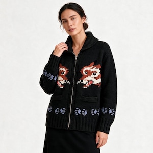 Puede incluir: Una chaqueta de punto negra con cremallera, cuello chal y dos bolsillos delanteros. La prenda presenta un patrón gráfico de dos cabezas de zorro estilizadas en marrón, blanco y rojo, con un motivo repetitivo de huellas azul claro en el dobladillo.