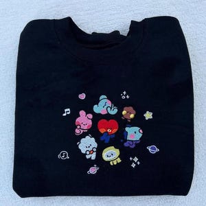 May include: Black sweatshirt with an embroidered design of cartoon characters from the popular K-pop group BTS. The characters are surrounded by stars and musical notes.