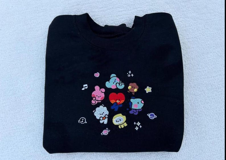May include: Black sweatshirt with an embroidered design of cartoon characters from the popular K-pop group BTS. The characters are surrounded by stars and musical notes.