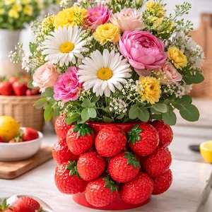 Op de afbeelding: Een levendig bloemstuk in een rode vaas in de vorm van een aardbei. Het boeket bevat madeliefjes, roze rozen, gele ranonkels en andere kleurrijke bloemen. Verse aardbeien en citroenen zijn ook zichtbaar.