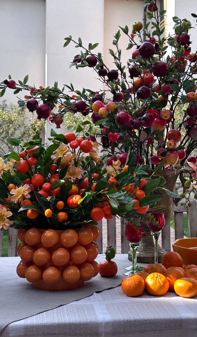 K&ouml;nnte beinhalten: Eine Tischdekoration mit zwei orangefarbenen Keramikvasen, gef&uuml;llt mit Orangen und roten Beeren. Die Vasen stehen auf einer wei&szlig;en Tischdecke mit einem karierten Muster. Auf dem Tisch stehen auch zwei Weingl&auml;ser und eine Sch&uuml;ssel mit Orangen.
