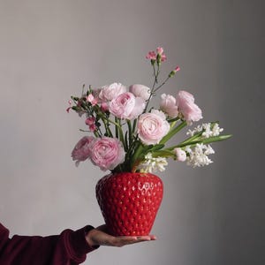 Könnte beinhalten: Eine leuchtend rote Keramikvase in Erdbeerform, gefüllt mit einem Blumenarrangement aus rosa Blüten und weißen Hyazinthen. Die Vase hat eine strukturierte Oberfläche. Das Arrangement wird in der Hand einer Person gehalten und bildet einen schönen Kontrast.