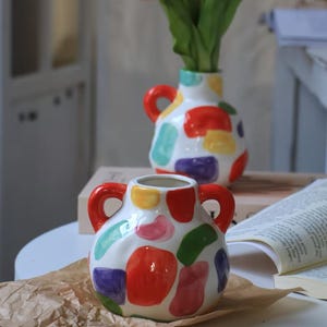 Puede incluir: Dos jarrones de cerámica blanca con asas rojas y diseños abstractos coloridos. Los jarrones están decorados con formas rojas, amarillas, moradas, verdes y rosas. Un jarrón contiene hojas verdes. Los jarrones están sobre una mesa blanca.