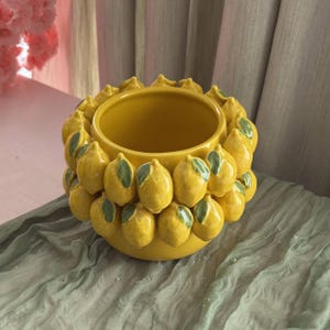Può includere: Un vaso di ceramica giallo decorato con limoni con foglie verdi. Il vaso ha un'ampia apertura ed è posto su un tessuto verde. Lo sfondo presenta elementi rosa e beige.