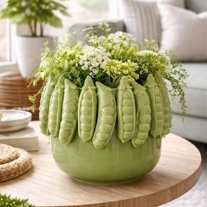 May include: A green ceramic vase with a pea pod design around the rim, filled with green and white flowers. The vase sits on a round wooden table. A plant in a white pot is in the background.