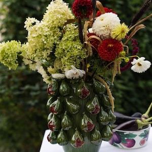 May include: A green ceramic vase with a vertical ribbed texture and three-dimensional fig decorations, filled with a large bouquet of green hydrangeas, deep red dahlias, and white flowers. A matching bowl with fig illustrations sits nearby.