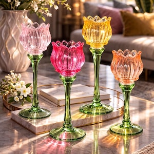 Op de afbeelding: Vier kleurrijke glazen kandelaars in roze, geel en oranje. Elke houder heeft een tulpvormige beker en een slanke groene steel. De kandelaars zijn geplaatst op een tafel met marmeren blad, die licht reflecteert.