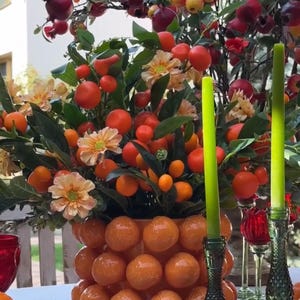 K&ouml;nnte beinhalten: Eine Keramikvase in Form einer Orangensammlung, gef&uuml;llt mit einem Blumenarrangement aus kleinen orangen Fr&uuml;chten, gr&uuml;nen Bl&auml;ttern und pfirsichfarbenen Bl&uuml;ten. Zwei gr&uuml;ne Kerzenhalter aus Glas mit hohen gr&uuml;nen Kerzen stehen auf einer wei&szlig;en Tischdecke.