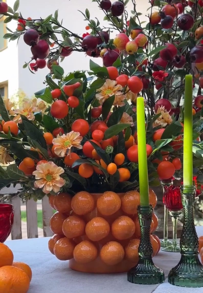 K&ouml;nnte beinhalten: Eine Keramikvase in Form einer Orangensammlung, gef&uuml;llt mit einem Blumenarrangement aus kleinen orangen Fr&uuml;chten, gr&uuml;nen Bl&auml;ttern und pfirsichfarbenen Bl&uuml;ten. Zwei gr&uuml;ne Kerzenhalter aus Glas mit hohen gr&uuml;nen Kerzen stehen auf einer wei&szlig;en Tischdecke.
