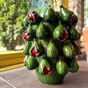 Puede incluir: Un jarrón cilíndrico de cerámica verde con una textura acanalada vertical y numerosas esculturas de higos en tres dimensiones adheridas al exterior. Los higos son de color verde brillante con interiores expuestos que muestran una pulpa roja.