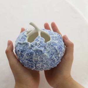 Op de afbeelding: Een witte keramische vaas met een blauw bloemenontwerp. De vaas is gevormd als een hortensia-bloem en heeft een kleine opening aan de bovenkant.