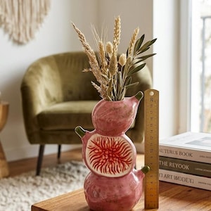 Peut inclure: Un vase en céramique rose en forme de figue empilée avec une section transversale montrant des détails intérieurs rouges. Une règle en bois placée à côté du vase indique une hauteur d'environ 19 cm. Le vase contient du blé séché et du feuillage vert.