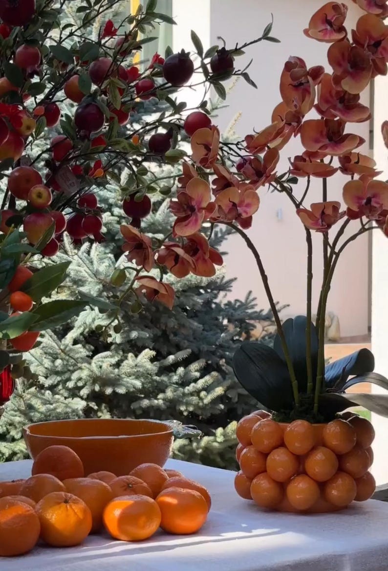 K&ouml;nnte beinhalten: Ein wei&szlig;er Tisch mit einer Sch&uuml;ssel Orangen und einem dekorativen orangen Topf mit einer rosa Orchideenpflanze. Die Orchideenpflanze hat mehrere rosa Bl&uuml;ten.