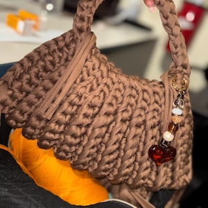 May include: A brown crocheted handbag featuring a thick braided handle and a textured stitch pattern. A decorative charm hangs from a gold-toned ring on the side, consisting of several small beads and a prominent dark red heart-shaped pendant.