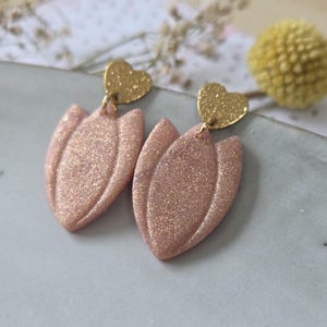May include: A pair of drop earrings featuring a gold-coloured heart-shaped stud at the top. Attached to each heart is a pale pink, glittery, petal-shaped pendant with a raised central ridge, creating a layered, textured appearance in the design.