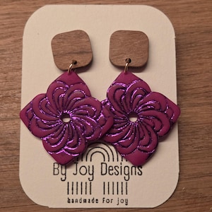Op de afbeelding: Handgemaakte oorbellen met vierkante houten studs en magenta bloemvormige hangers. De hangers hebben een metallic glans en een gedetailleerd, wervelend patroon. De oorbellen worden gepresenteerd op een kaart met de tekst "By Joy Designs" en "handmade for joy."