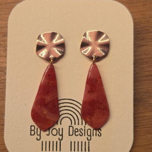 Puede incluir: Un par de pendientes hechos a mano con la parte superior de color cobre y la parte inferior roja en forma de lágrima con motas doradas. Los pendientes se muestran en una tarjeta que dice "By Joy Designs" y "handmade for joy".