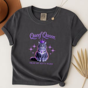 May include: Dark gray t-shirt featuring a regal cat illustration wearing a crown and necklace. The text "Queef Queen" is above the cat, with "HEAR MY KITTY PURR" below. The design includes purple stars.