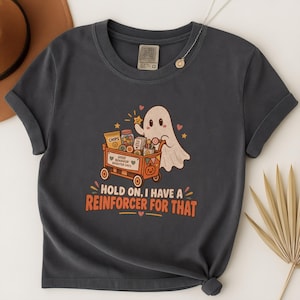May include: A dark gray t-shirt featuring a graphic of a white ghost pushing an orange cart filled with snacks and toys. The cart is labeled "GOOD BEHAVIOR BRIGHTER DAYS" and the text below reads "HOLD ON, I HAVE A REINFORCER FOR THAT" in orange.