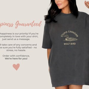 May include: Dark grey t-shirt with the text "STUPID CANADIAN WOLF BIRD" and a graphic of a loon. The shirt also has the text "Happiness Guaranteed" and other text. The shirt is a casual, oversized fit.