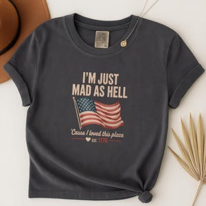 May include: Dark gray t-shirt with the text "I'M JUST MAD AS HELL" above an American flag graphic. Below the flag, the text reads "'Cause I loved this place" and "EST. 1776". The shirt has rolled-up sleeves.