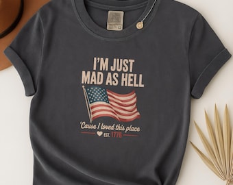 May include: Dark gray t-shirt with the text "I'M JUST MAD AS HELL" above an American flag graphic. Below the flag, the text reads "'Cause I loved this place" and "EST. 1776". The shirt has rolled-up sleeves.