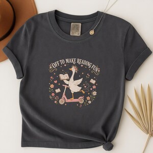 May include: A dark gray short-sleeve t-shirt featuring a graphic of a white goose wearing a flower crown, riding a pink scooter, and holding an open book. The text "OFF TO MAKE READING FUN" is printed above the goose, surrounded by small flowers.
