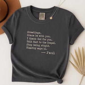 May include: A dark charcoal gray short-sleeve t-shirt featuring white typewriter-style text that reads: Greetings, Grace be with you. I thank God for you. Hold fast to the Gospel. Stop being stupid. Timothy says hi. — Paul. The shirt has a crew neckline.
