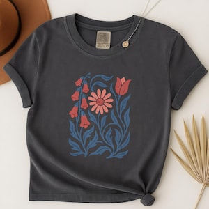 May include: Dark gray t-shirt featuring a floral design with red, pink, and blue elements. The graphic includes a central daisy, bell-shaped flowers, and a tulip, all set against blue foliage. The shirt has short sleeves and a crew neck.