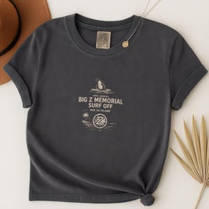 May include: Dark gray t-shirt with rolled sleeves featuring a graphic for the "Big Z Memorial Surf Off" with a penguin on a surfboard. The text includes "Pen Gu Island" and a logo. A necklace with a pendant is visible.