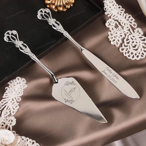 May include: Silver wedding cake knife and server set with ornate handles. The server is inscribed with "Kathy & John June 23, 2023" and the knife is inscribed with "A & E Archer & Jillian 17 March 2023".