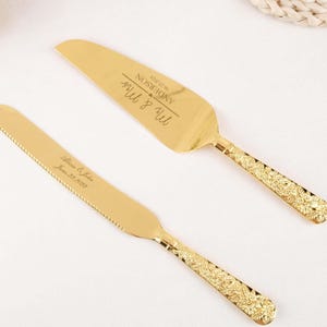 May include: A gold cake knife and server set. The server is engraved with "Mr & Mrs" and a name and date. The knife is engraved with a name and date. Both have decorative handles.