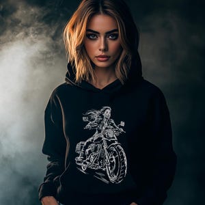 Puede incluir: Sudadera con capucha negra con un dibujo lineal blanco de una persona en una motocicleta. El diseño muestra al motorista con una chaqueta y pelo suelto. El fondo es gris oscuro con un efecto ahumado. La sudadera tiene capucha.